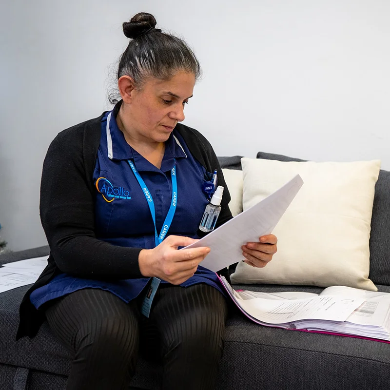Apollo Care carer in branded blue uniform and lanyard reviewing a client's care-plan paperwork on a home sofa during a domiciliary visit