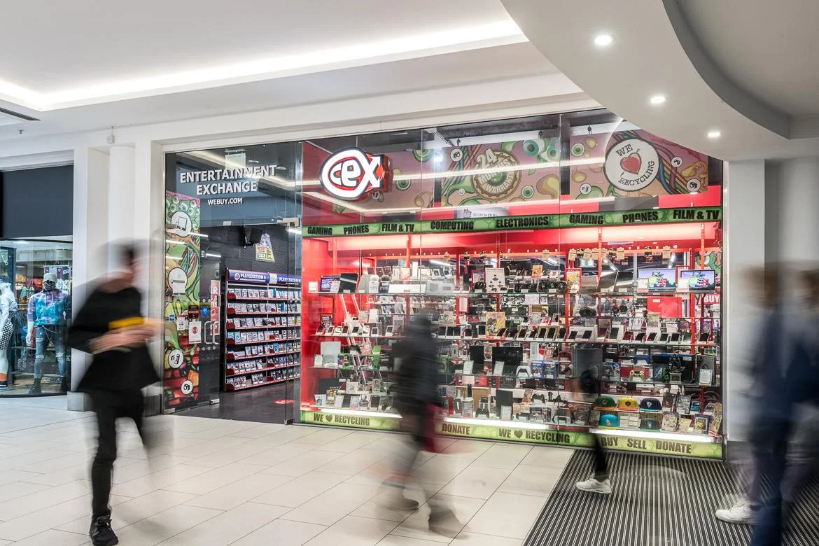 CeX shopping-mall shopfront with red 'Entertainment Exchange' signage, 'WeBuy.com' branding and product departments for Gaming, Phones, Film and TV, Computing and Electronics on display inside