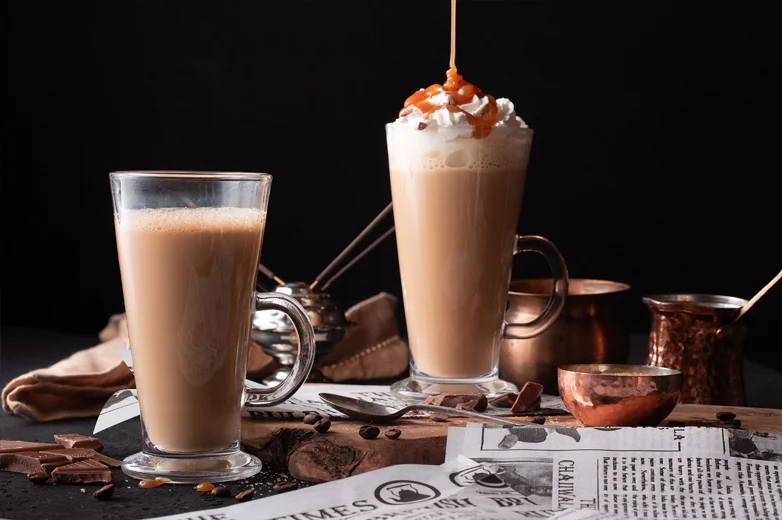 Chaiiwala signature karak chai latte in tall glass mugs - one with whipped cream and caramel drizzle, served alongside traditional copper chai pots