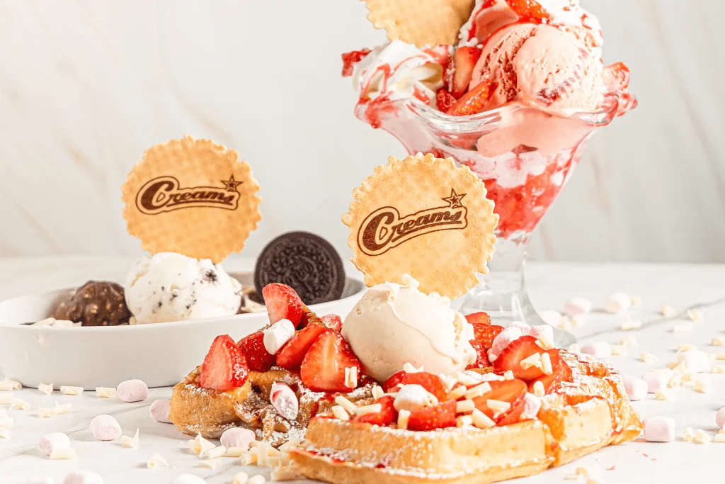 Creams Cafe close-up plate: a Belgian waffle topped with fresh strawberries, vanilla ice cream and cream, paired with a strawberry-gelato sundae glass, Oreo crumb plate and branded Creams wafers