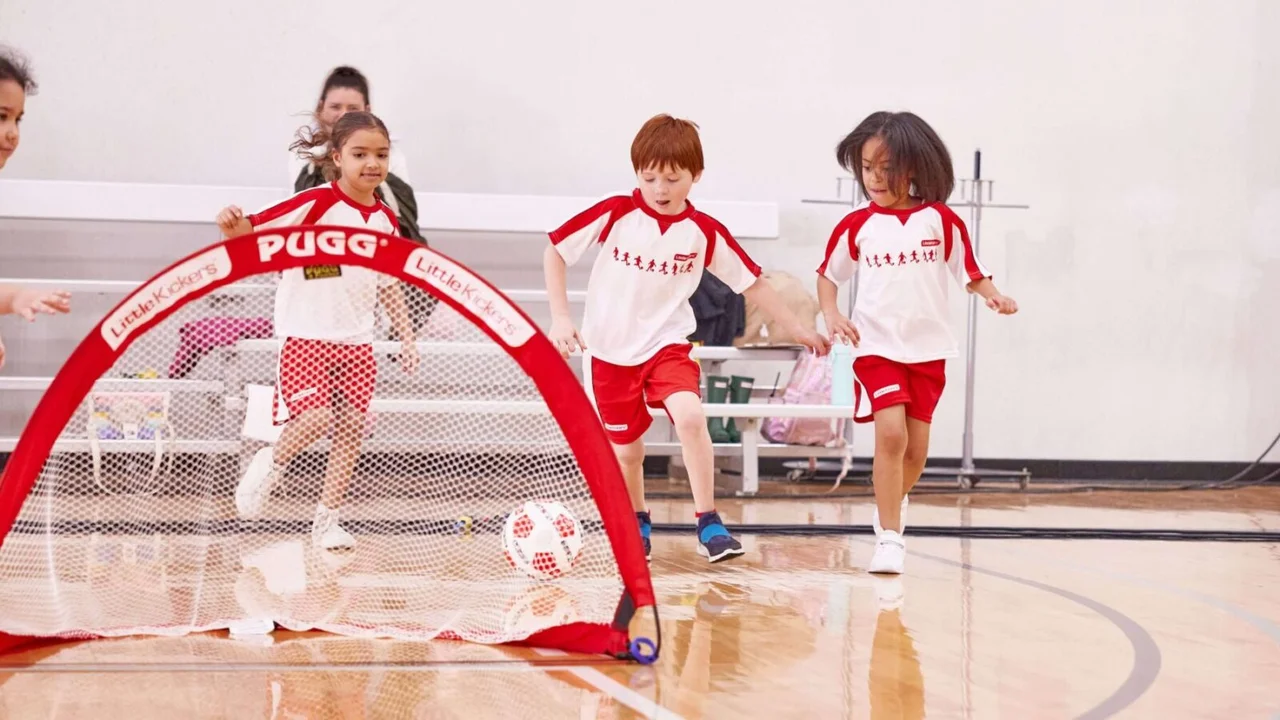 Young children in red and white Little Kickers kit playing a football game indoors with a pop-up PUGG goal in a school sports hall