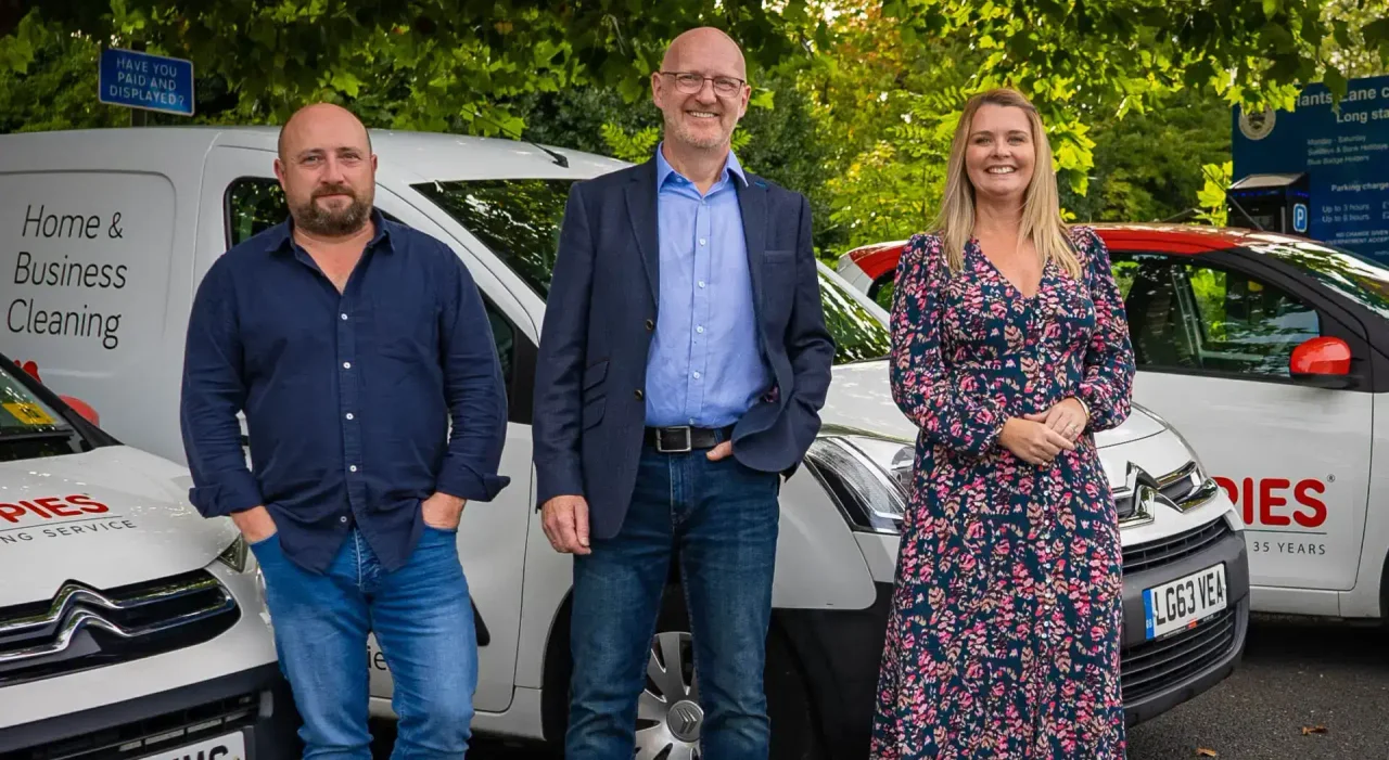 Three Poppies team members - two men in business-casual attire and a woman in a floral dress - standing in front of branded white Poppies 'Home & Business Cleaning' vans on a tree-lined street