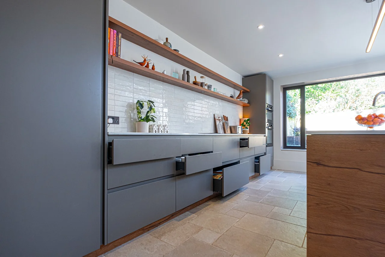 Raison Home UK modern grey handleless kitchen with open walnut shelving, white metro tile splashback, integrated appliances and a stone-tiled floor