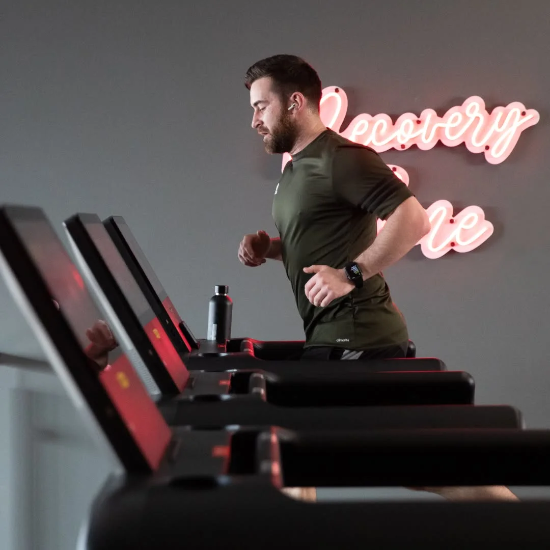 Male Snap Fitness member running on a treadmill in a green Adidas tee, with a pink neon 'Recovery' script sign glowing on the wall behind, illustrating the brand's modern cardio and recovery offering