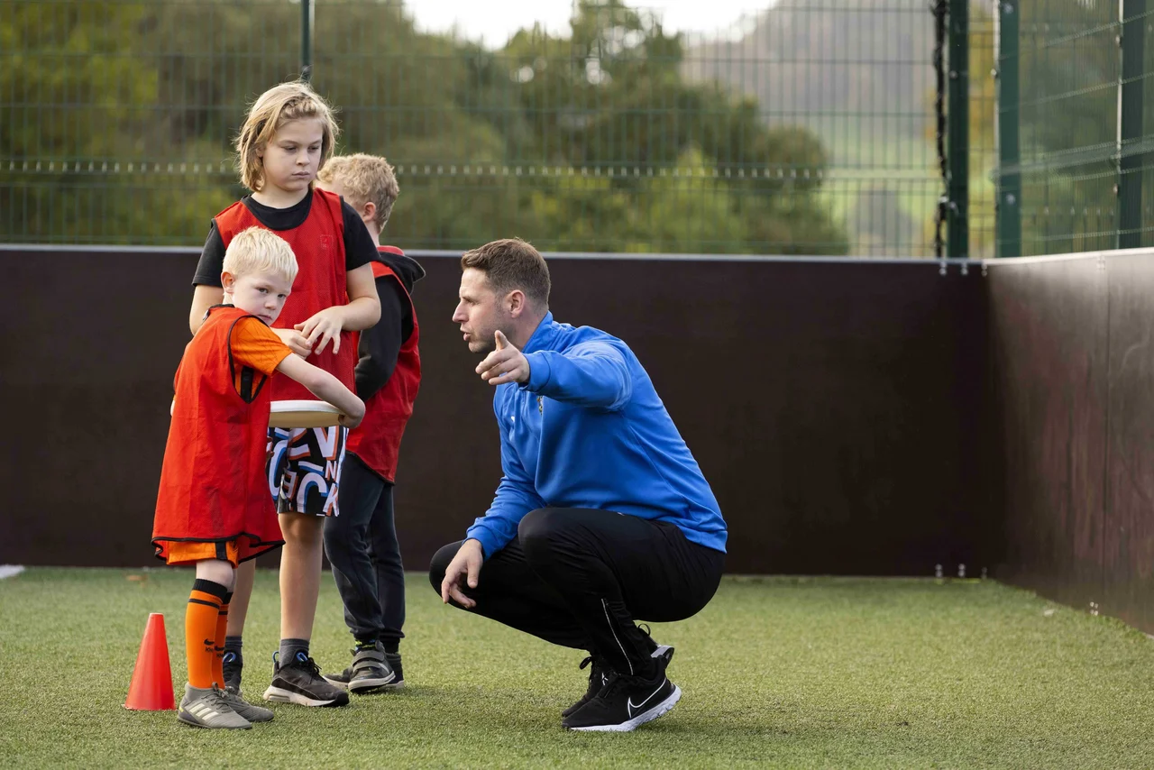 SportsCool coach in a blue branded jacket crouched on an astroturf pitch giving instructions to two children in orange and red SportsCool bibs, behind a metal mesh fence - a typical school-based SportsCool session