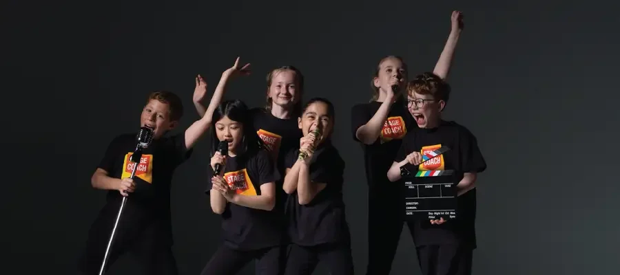 Six Stagecoach Performing Arts students in black branded tees performing animatedly with microphones and a film clapperboard against a dark grey backdrop - typical Saturday class energy