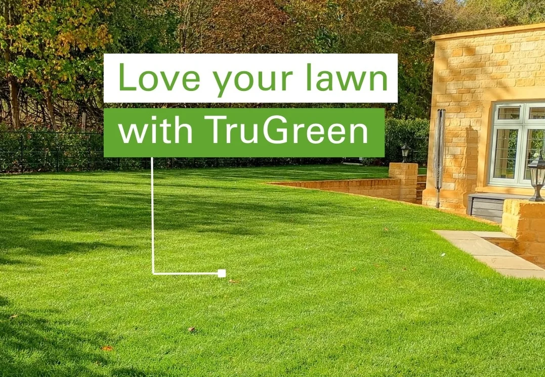 Beautifully manicured green lawn beside a Cotswold stone-built UK home, with a green-and-white marketing card reading 'Love your lawn with TruGreen' standing in the grass on a lawn-sign stake